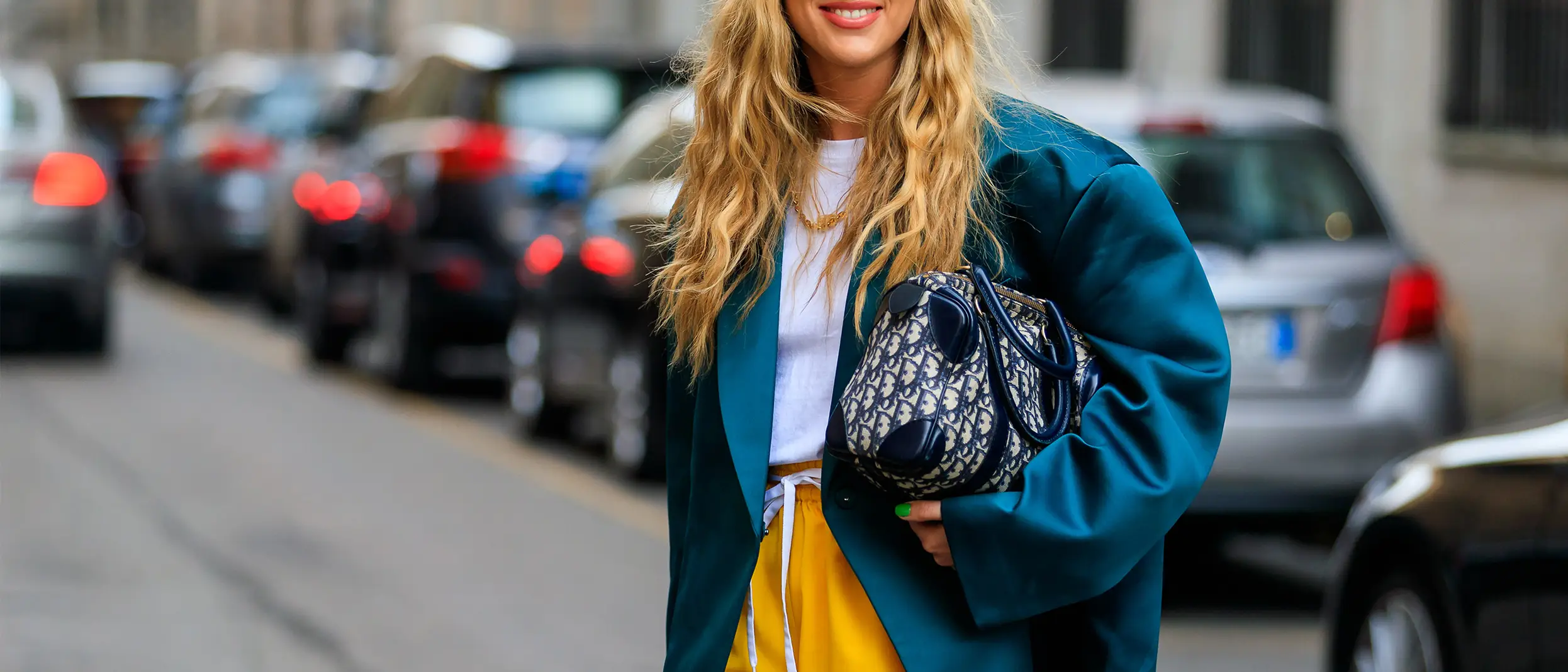Woman wearing a teal blazer, yellow skirt, and holding a Dior handbag, smiling on a city street. Woman wearing a teal blazer, yellow skirt, and holding a Dior handbag, smiling on a city street.